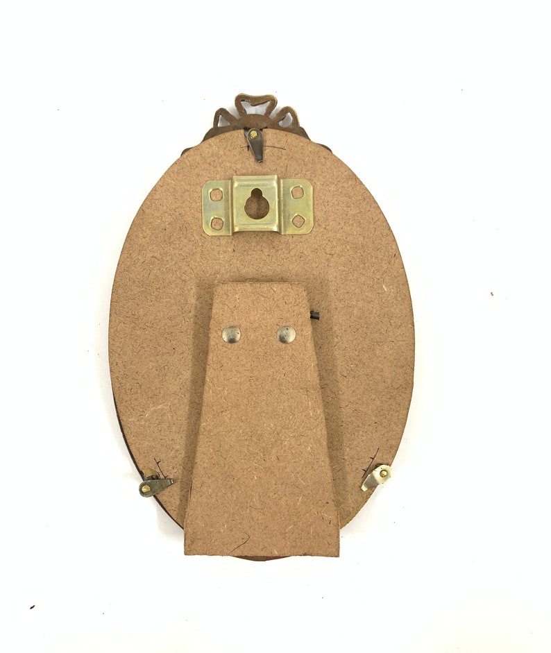 May include: Back of an oval-shaped picture frame with a decorative bronze-colored top. The frame has a cardboard backing with a metal hanger and a cardboard stand attached with small metal hinges and rivets.