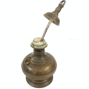 Puede incluir: Una botella de latón vintage con tapa y un largo pico delgado. La botella tiene un cuerpo redondeado y una pequeña asa. La botella está en buen estado, con algunas señales de desgaste.