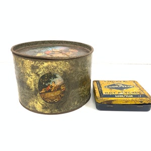 Può includere: Due scatole di latta vintage. La scatola più grande è rotonda e presenta un motivo floreale dorato e marrone con una scena di persone in un giardino. La scatola più piccola è rettangolare e presenta un design giallo e blu con il testo "Vulcanized Goodyear Hot Patches Goodyear".