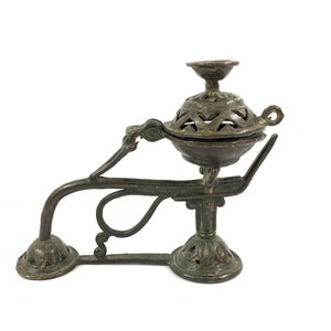 Op de afbeelding: Een bronzen wierookbrander met een decoratief, opengewerkt ontwerp. De brander heeft een ronde basis en een gebogen arm met een handvat. De brander staat op een witte achtergrond.