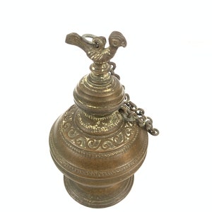 Puede incluir: Una lámpara de aceite de latón con un diseño ornamentado y un asa en forma de pájaro. La lámpara tiene una cadena unida al asa.