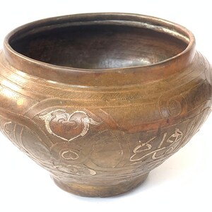 May include: A round, brass bowl with intricate engraved designs. The bowl has a wide, rounded base and a slightly flared rim. The engravings are in a silver color and feature geometric patterns and floral motifs.