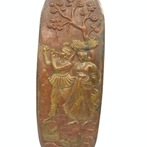May include: An oval-shaped copper wall hanging with a relief design of a couple standing under a tree. The man is playing a flute and the woman is carrying a basket on her head. The couple is dressed in traditional Indian clothing and the background is a stylized landscape.