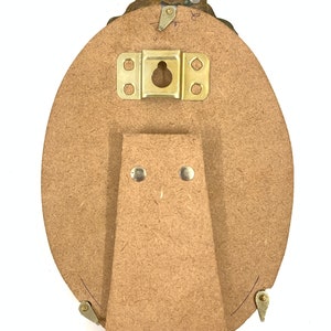 May include: Back of an oval picture frame with a decorative brass top. The frame has a wooden backing with a brass hanger and a wooden easel stand attached. The frame is designed to stand on a flat surface.