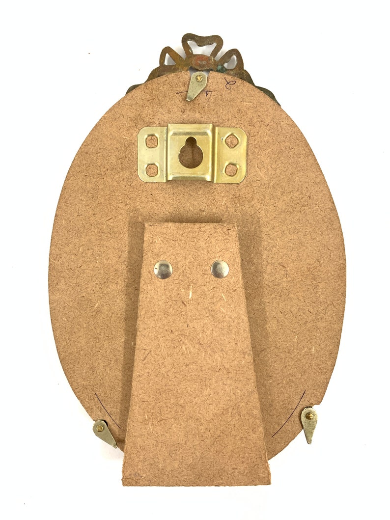 May include: Back of an oval picture frame with a decorative brass top. The frame has a wooden backing with a brass hanger and a wooden easel stand attached. The frame is designed to stand on a flat surface.