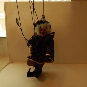 May include: A marionette puppet featuring a figure with white hair, a pointed hat, and a black cloak adorned with orange crescent moon and star patterns. The puppet is suspended by black strings and holds a small star-shaped wand in one hand.