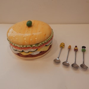 May include: A ceramic hamburger-shaped container with a green knob on top, and four decorative spoons. The container has a bun, lettuce, tomato, cheese, and patty design. The spoons have colorful food-themed handles.