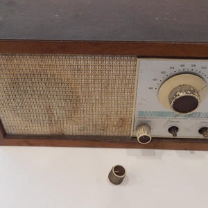 May include: Vintage wooden radio with a light-colored speaker cover and a cream-colored control panel. The radio has a large tuning dial and smaller knobs for volume, bass, and treble. A detached knob sits in front of the radio.