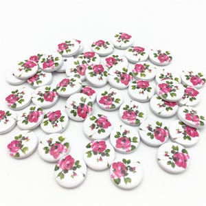 May include: A collection of round, white wooden buttons. Each button features a pink rose floral design with green leaves. The buttons are scattered across a white surface, showcasing their decorative floral pattern.