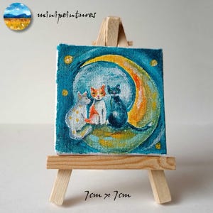 Puede incluir: Una pintura en miniatura sobre lienzo de tres gatos sentados en una luna creciente. La luna es amarilla y naranja, y el fondo es azul oscuro con estrellas blancas. La pintura mide 7 cm por 7 cm y se muestra en un pequeño caballete de madera.