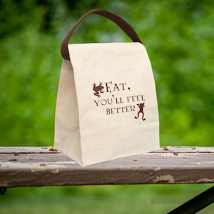 May include: A white canvas lunch bag with brown leather handles. The bag has a brown frog graphic and the text "EAT, YOU'LL FEEL BETTER".
