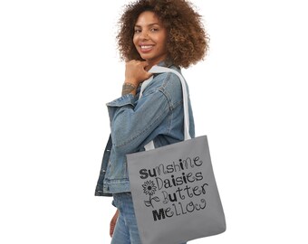 May include: A gray tote bag with white handles featuring the words "Sunshine Daisies Butter Mellow" in a whimsical font, along with a sunflower illustration. The bag is carried over the shoulder.