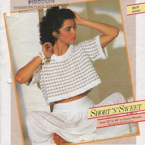 May include: A white crocheted top with a brown and white checkered pattern. The top is cropped and has short sleeves. The model is wearing a white skirt and is sitting down. The text "SHORT 'N' SWEET" is printed on a yellow banner. The text "Sizes 30 to 40" in Coton Mousse is printed below the banner. The text "See inside for alternative yarns" is printed below the size information.