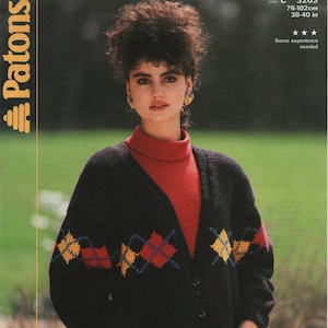 Może przedstawiać: Czarny kardigan z rombowym wzorem w kolorze czerwonym, żółtym i niebieskim. Kardigan ma kieszenie i zapięcie na guziki. Modelka ma na sobie pod spodem czerwony golf.