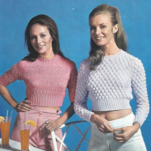 May include: Two women wearing cropped sweaters. The woman on the left is wearing a pink sweater with a lacy design and white pants. The woman on the right is wearing a light purple sweater with a textured design and white pants.