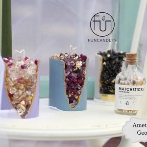 Peut inclure: Trois bougies en forme de g&eacute;ode d'am&eacute;thyste avec des accents dor&eacute;s et un flacon d'allumettes. Les bougies sont violettes, bleues et noires. Le flacon d'allumettes indique "Matchsticks Hand Made". Le texte "Am&eacute;thyste Geode" est &eacute;crit sous les bougies.