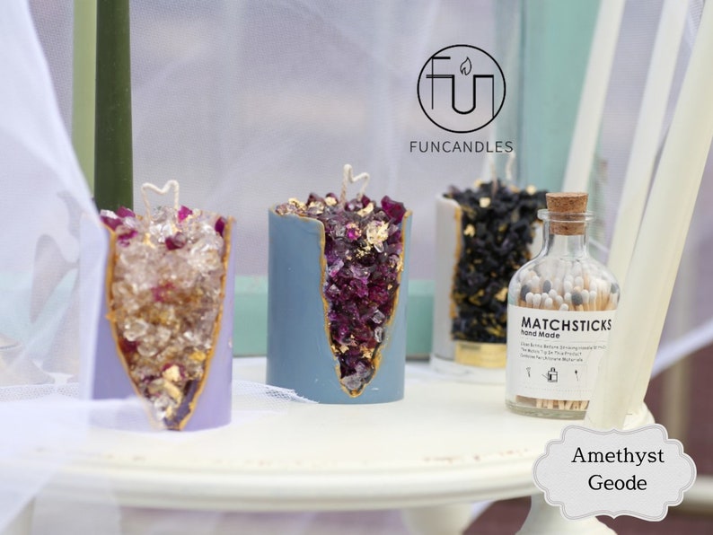 Peut inclure: Trois bougies en forme de g&eacute;ode d'am&eacute;thyste avec des accents dor&eacute;s et un flacon d'allumettes. Les bougies sont violettes, bleues et noires. Le flacon d'allumettes indique "Matchsticks Hand Made". Le texte "Am&eacute;thyste Geode" est &eacute;crit sous les bougies.