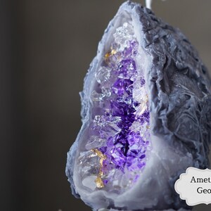 Pu&ograve; includere: Una candela a forma di geode di ametista viola con un esterno grigio e accenti dorati. La candela ha la forma di una semisfera e presenta una superficie testurizzata dettagliata.