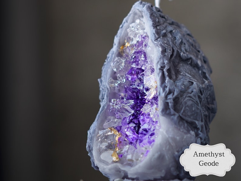 Pu&ograve; includere: Una candela a forma di geode di ametista viola con un esterno grigio e accenti dorati. La candela ha la forma di una semisfera e presenta una superficie testurizzata dettagliata.
