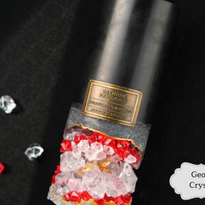 May include: A black candle with a gold label that reads "Premium Fragrance, Perfumery Goffret, Made in Belgium". The candle is surrounded by clear and red crystals, resembling a geode. The label is partially obscured by the crystals. The text "Geode Crystal" is written in a white label with a decorative border.