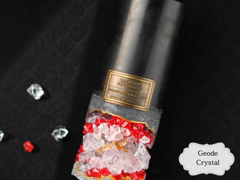 May include: A black candle with a gold label that reads "Premium Fragrance, Perfumery Goffret, Made in Belgium". The candle is surrounded by clear and red crystals, resembling a geode. The label is partially obscured by the crystals. The text "Geode Crystal" is written in a white label with a decorative border.