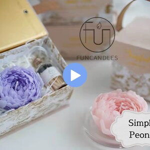 May include: A purple peony candle in a white box with a small bottle of essential oil. The box is decorated with a gold and white pattern. The candle is a beautiful and unique gift for any occasion.