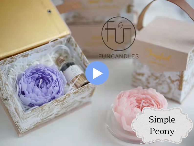 May include: A purple peony candle in a white box with a small bottle of essential oil. The box is decorated with a gold and white pattern. The candle is a beautiful and unique gift for any occasion.