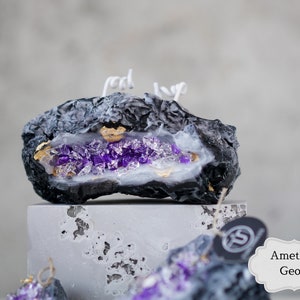 Pu&ograve; includere: Candela a forma di geode di ametista nera e viola con accenti dorati. La candela ha la forma di una roccia e un interno bianco con cristalli viola.