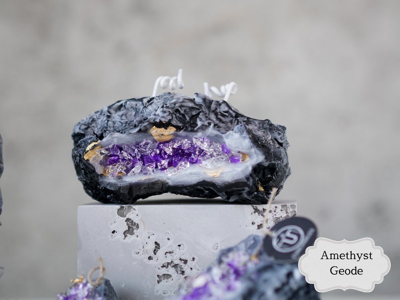 Pu&ograve; includere: Candela a forma di geode di ametista nera e viola con accenti dorati. La candela ha la forma di una roccia e un interno bianco con cristalli viola.