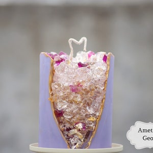 Peut inclure: Bougie violette avec un bord dor&eacute; et un centre transparent rempli de cristaux clairs et roses. La bougie est &eacute;tiquet&eacute;e "Am&eacute;thyste G&eacute;ode".