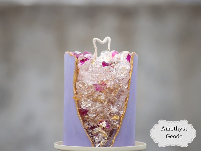 Peut inclure: Bougie violette avec un bord dor&eacute; et un centre transparent rempli de cristaux clairs et roses. La bougie est &eacute;tiquet&eacute;e "Am&eacute;thyste G&eacute;ode".