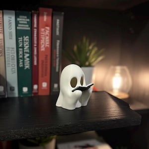 Pode incluir: Uma estatueta de fantasma branco lendo um pequeno livro preto, colocada em uma prateleira preta. O fantasma tem grandes buracos redondos para os olhos e segura o livro com as mãos. Livros e uma pequena planta são visíveis ao fundo.