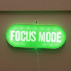 May include: A green, illuminated sign with the words "FOCUS MODE" in white lettering. The sign is rectangular with rounded edges and is mounted on a light-colored wall. A white cord extends from the bottom of the sign.