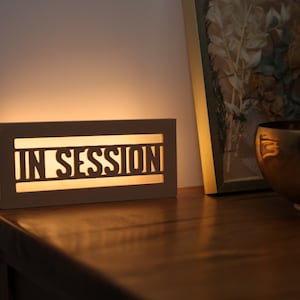 May include: A rectangular, illuminated sign with the words "IN SESSION" cut out. The sign is made of a light-colored material and emits a warm glow. A white cord extends from the sign. The sign is on a wooden surface.
