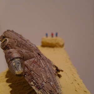 Puede incluir: Un modelo marrón y gris del  Millennium Falcon  parcialmente enterrado en la arena. La nave espacial está inclinada hacia un lado y tiene muchos detalles.