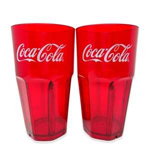 May include: Two translucent red plastic tumblers featuring the white Coca-Cola logo printed on the front. Each cup has a tapered shape with vertical ribbed detailing along the lower sides and a flat base, designed for holding cold beverages.