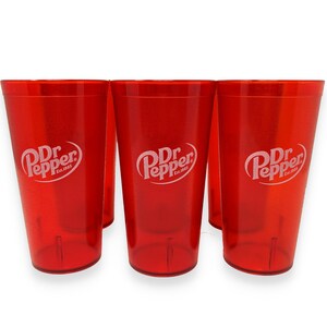 May include: Three translucent red plastic tumblers, each featuring the white Dr Pepper logo with the text Est.1885. The cups have a tapered cylindrical shape with a smooth finish and a slightly reinforced base, displaying a consistent vibrant red color.