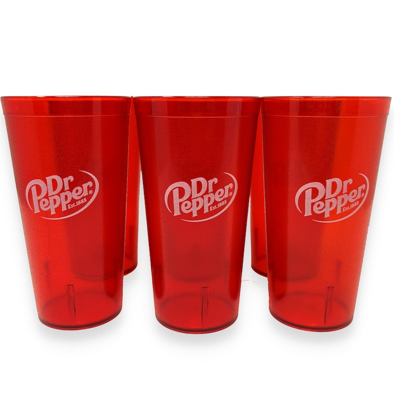 May include: Three translucent red plastic tumblers, each featuring the white Dr Pepper logo with the text Est.1885. The cups have a tapered cylindrical shape with a smooth finish and a slightly reinforced base, displaying a consistent vibrant red color.