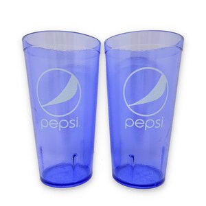 May include: Two translucent blue plastic tumblers featuring the white Pepsi logo and text printed on the front. Each cup has a textured surface and a tapered cylindrical shape with a flat base, designed for serving cold beverages.