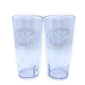 May include: Two clear, cylindrical plastic tumblers featuring the Dr Pepper logo etched on the front. Each glass displays the text Dr Pepper Est. 1885 within an oval frame. The tumblers have a textured base and a smooth, transparent body design.