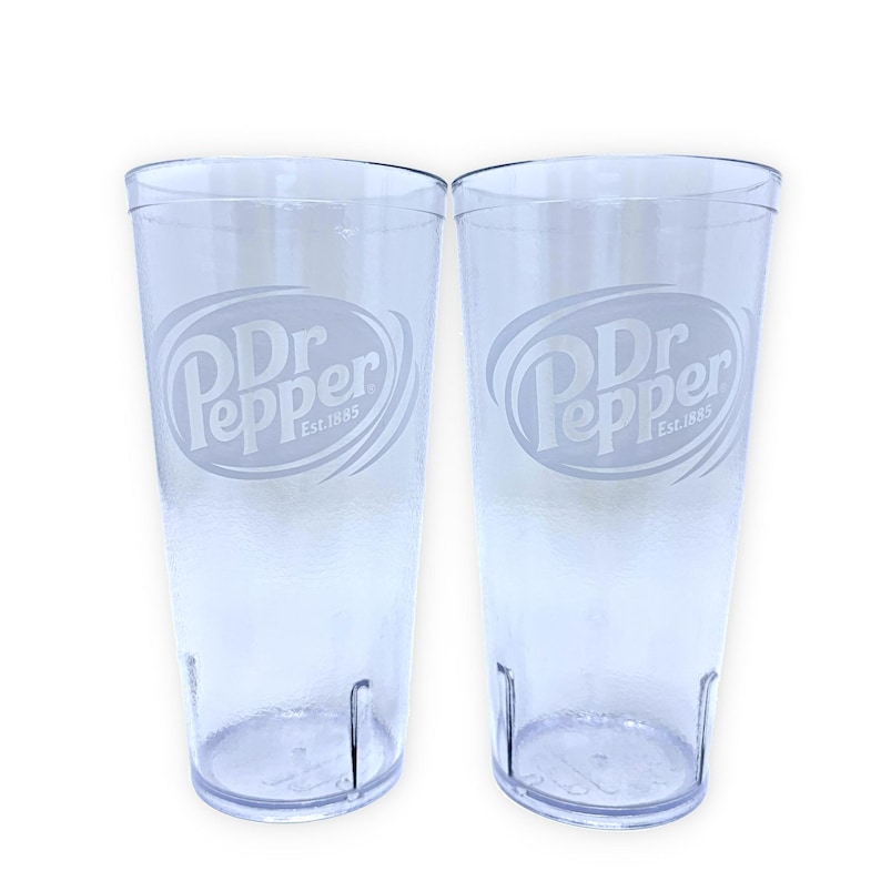 May include: Two clear, cylindrical plastic tumblers featuring the Dr Pepper logo etched on the front. Each glass displays the text Dr Pepper Est. 1885 within an oval frame. The tumblers have a textured base and a smooth, transparent body design.