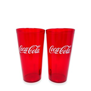 May include: Two identical translucent red plastic tumblers featuring the white Coca-Cola logo printed on the front. Each cup has a tapered cylindrical shape with a smooth finish and a flat base, designed for holding cold beverages in a kitchen setting.