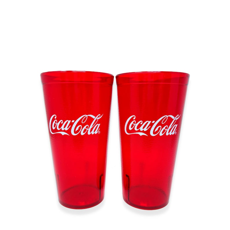 May include: Two identical translucent red plastic tumblers featuring the white Coca-Cola logo printed on the front. Each cup has a tapered cylindrical shape with a smooth finish and a flat base, designed for holding cold beverages in a kitchen setting.