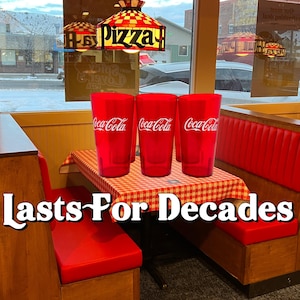 May include: Three red Coca-Cola branded plastic cups on a checkered tablecloth in a restaurant booth. The booth has red seating and a wooden table. The text "Lasts For Decades" is at the bottom of the image.