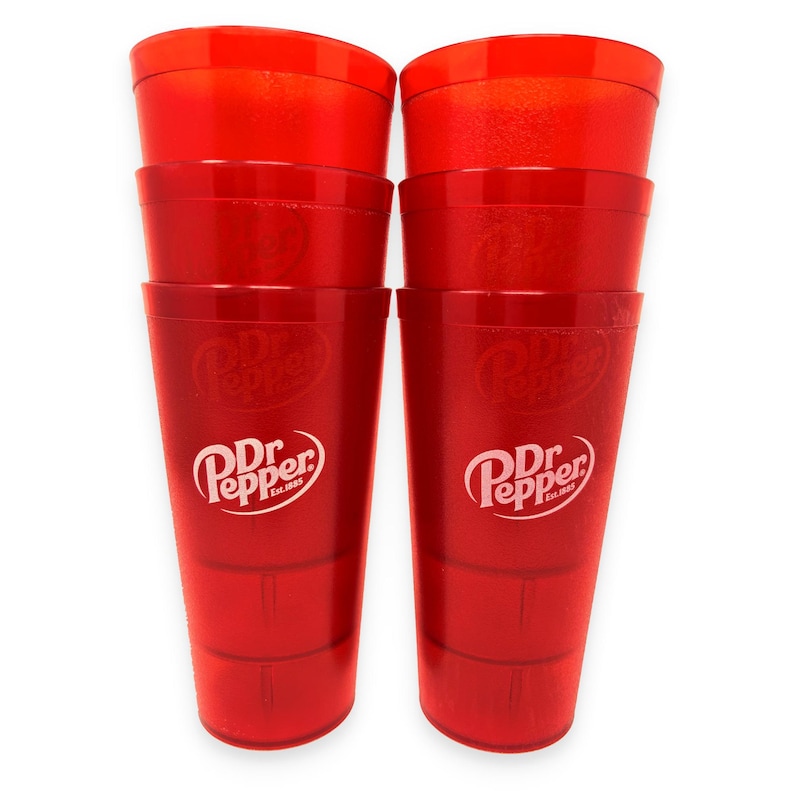 May include: A set of six stackable, translucent red plastic cups arranged in two columns of three. Each cup features the white Dr Pepper logo with the text Est. 1885 printed on the front. The cups have a tapered cylindrical shape with a smooth finish.