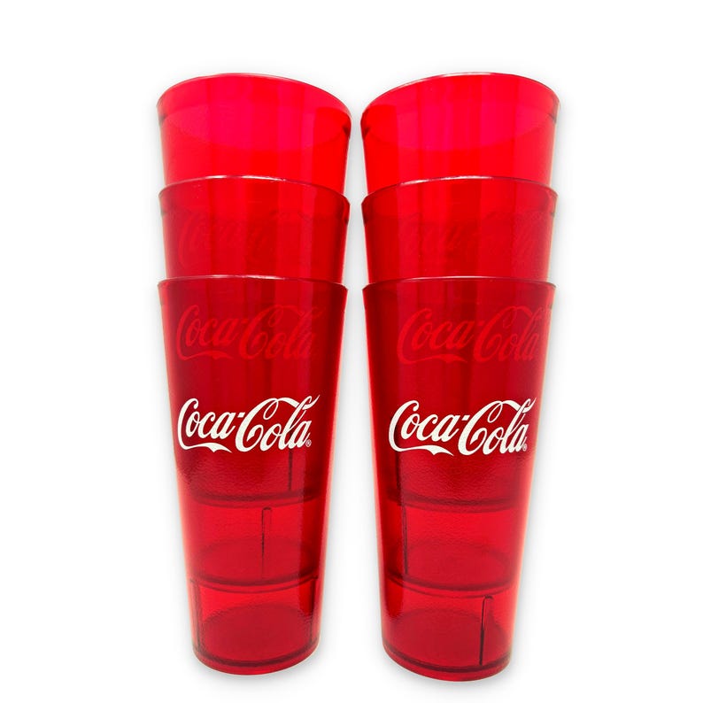 May include: Six red plastic Coca-Cola glasses stacked in two columns of three. Each glass has the Coca-Cola logo in white. The glasses are transparent and appear to be empty.