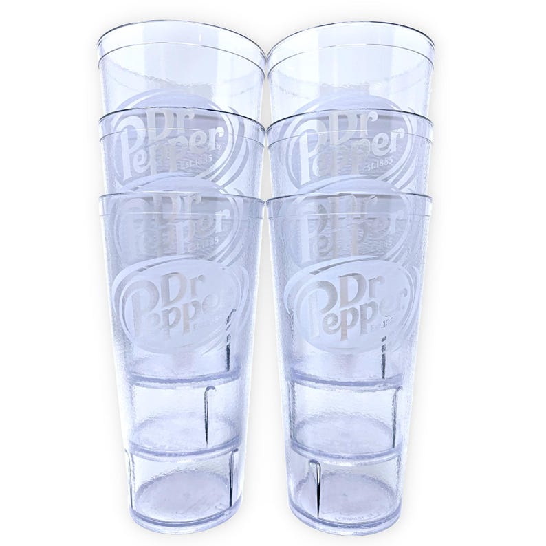 May include: A set of six clear plastic tumblers stacked in two columns of three. Each tumbler features a textured surface and a white etched Dr Pepper logo with the text Est. 1885. The cups have a tapered cylindrical shape with a smooth rim.