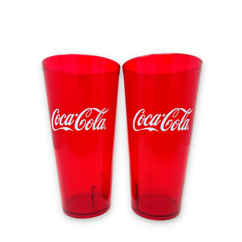 May include: Two tall, translucent red drinking glasses made of plastic. Each glass features the white Coca-Cola logo printed clearly on the front. The tumblers have a tapered cylindrical shape with a smooth finish and a flat base.