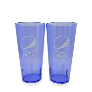 May include: Two tall, translucent purple plastic tumblers featuring a textured exterior surface. Each cup displays the white Pepsi logo and text printed on the front, with a tapered cylindrical shape and a reinforced base for stability.
