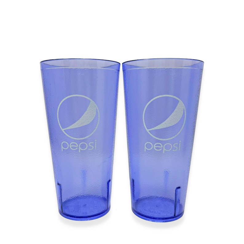 May include: Two tall, translucent purple plastic tumblers featuring a textured exterior surface. Each cup displays the white Pepsi logo and text printed on the front, with a tapered cylindrical shape and a reinforced base for stability.